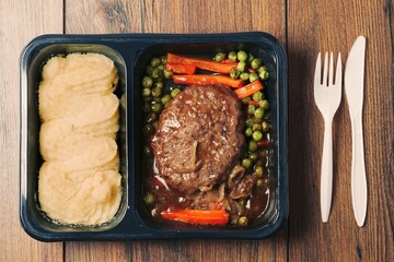 A meal consisting of burger, mashed potatoes and vegetables in plastic tray is served on wooden...