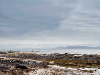 A beach with a rocky shoreline and a calm ocean. The sky is cloudy and the water is a deep blue. Galway bay, Ireland. Burren mountains in the background. Travel and tourism. Irish landscape.