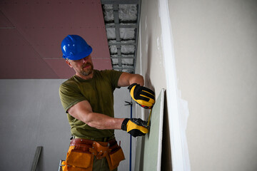 Construction worker wearing tool belt and hard hat measuring plasterboard using tape measure