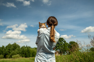 Obraz premium Domestic cat on leash walking outside for the first time, sitting on shoulder of loving woman owner, interested in new smells, worried and afraid, looking around. Walk with pets in nature. 