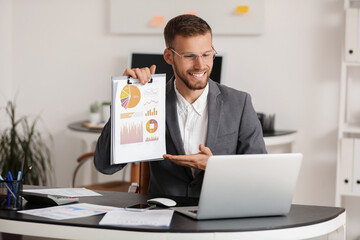 Young businessman in eyeglasses showing documents with graphs to laptop on video call in office