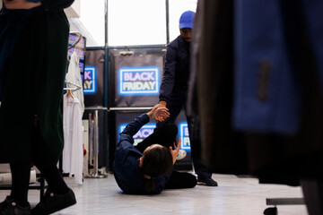 African American male security guard helping fallen injured man while controlling shop entrance during Black Friday sales. Shoppers hit stores in hunt for deals. Shopping madness concept