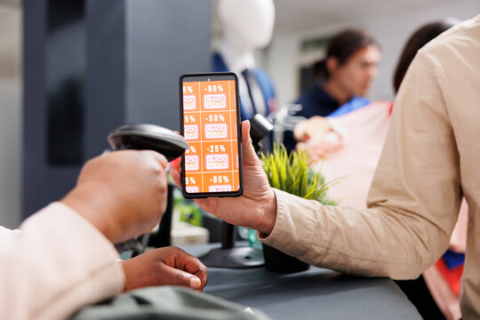 Digital coupon marketing. Customer using mobile discount code while shopping in clothing store on Black Friday, close up of man showing smartphone with digital voucher to retail employee at checkout