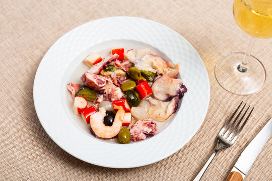 There is portion of seafood tapas salpicon on plate. Dish is made up of chopped octopus, squid, crab sticks, green and black olives, marinated cucumbers. Served with glass of chilled beer