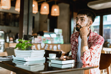 A bearded Asian man wearing glasses is thinking while reading a book at a cafe.