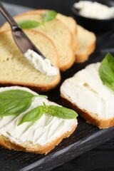 Pieces of bread with cream cheese and basil leaves on black table, closeup