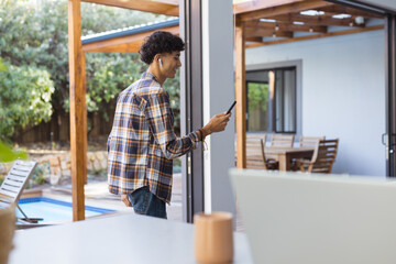 Using smartphone and wearing earbuds, teenage boy walking through modern home patio