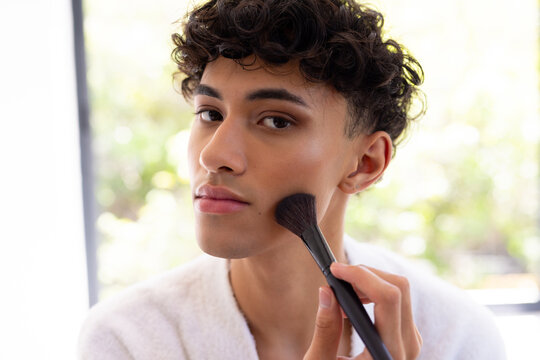Applying makeup, young man using brush for contouring at home, focusing on face