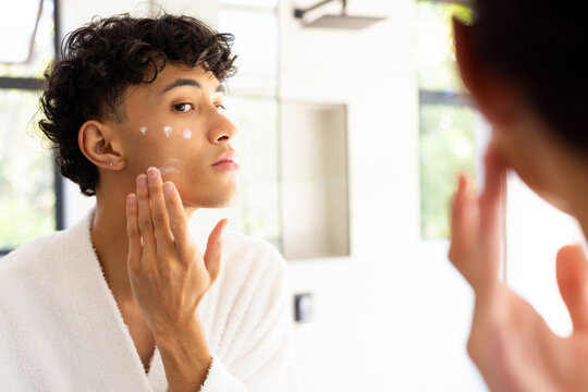Applying skincare cream, teenage boy in white robe looking in mirror