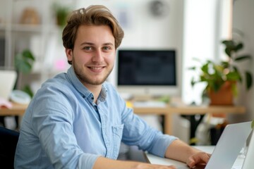 Fototapeta premium Portrait of a successful and confident young man in a blue shirt sitting in the office at a desk, working on a laptop and smiling at the camera 