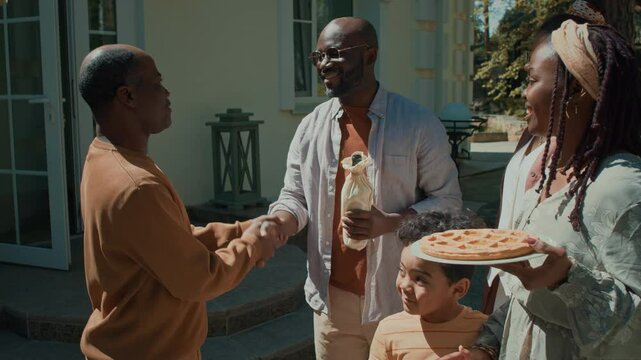 Medium shot of 30-year-old African American man greeting and shaking hands with father or male friend, holding bottle of wine, while visiting with wife and kids on national holiday in summer