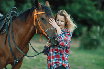 Obraz premium Happy blonde with horse in forest. Woman and a horse walking through the field during the day. Dressed in a plaid shirt and black leggings.