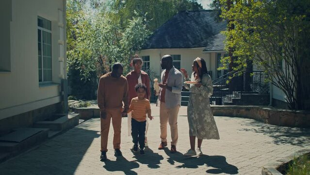 Full follow shot of multigenerational African American family walking on pavement in courtyard of suburban home, carrying cake and wine, on way to visit friends or relatives on summer national holiday