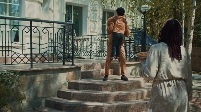 Full follow shot of young African American man and woman with young son walking to relatives home on visit, happy black uncle or granddad hugging nephew or grandson on front porch
