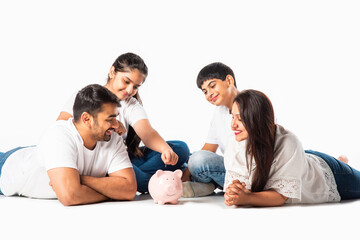 Indian asian young parents family with two kids saving or putting money in piggy bank lying on floor