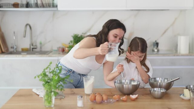 Happy mother and her daughter adding milk in bowl and whisking ingredients using metal corolla in the kitchen