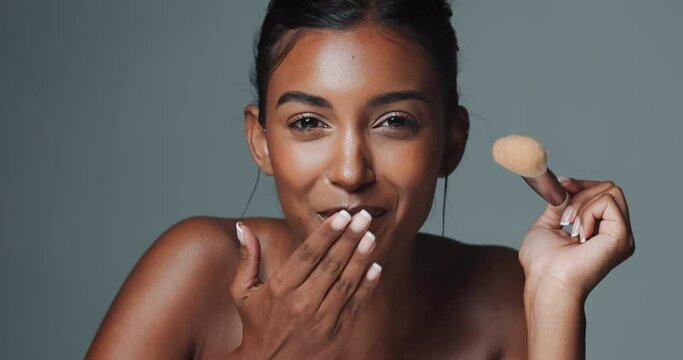 Kiss, makeup and face of Indian woman with brush, beauty treatment and wellness in studio. Female model, application and flirt blow with cosmetics, foundation and powder by gray background with pride