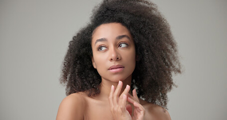 Beauty and healthcare concept - beautiful African American woman with curly afro hairstyle and clean, healthy skin touches her cheek and face with her hand, posing and looking at the camera