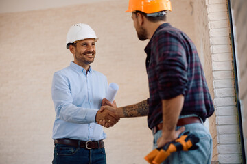 Happy building contractor shaking hands with  worker at construction site.