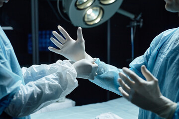 Side view of unrecognizable surgeon putting on gloves in operating room with medical assistant...