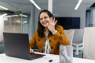 Obraz premium A young, beautiful woman worker in an office who is waving in front of her computer screen, with her hand outstretched and smiling in a yellow coat