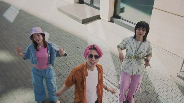High angle shot of pink haired Asian boy with two pretty girls standing slightly behind dancing to k-pop music outdoors on pavement under sunlight