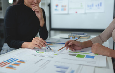 Two businesswomen meet, discuss, brainstorm, point at graphs and charts to analyze online market data, calculate balance sheets, accounts, net profit to plan company sales strategies.