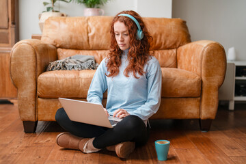 Young Woman With Red Hair Using Laptop While Wearing Headphones and Sitting on Floor
