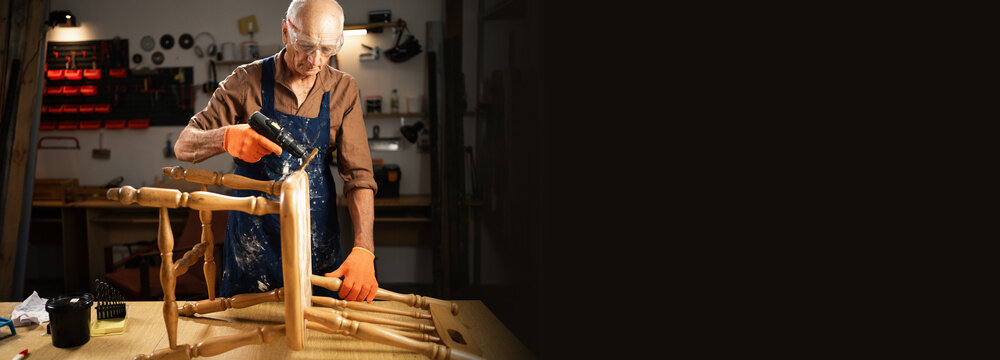 An elderly man in a workshop wearing protective glasses and orange gloves inserts and sands an old chair before restoration.