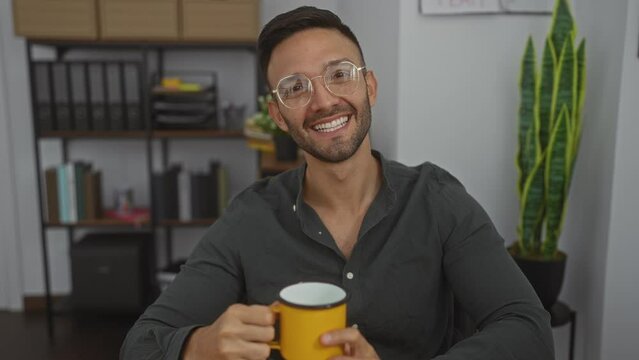 Young man smiling and waving at the camera while holding a yellow mug in an office environment filled with bookshelves and plants