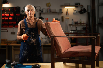 An elderly man in orange gloves and a blue apron begins restoring an old chair in a workshop. He...
