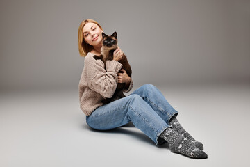 A woman with short hair sits on the floor, cradling a cat in her arms with a peaceful expression.