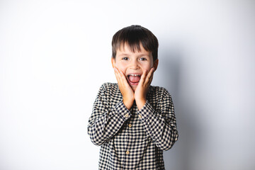 Portrait of funny young boy looking at camera with open mouth and big eyes, hands on cheek, shocked surprised expression against white background.