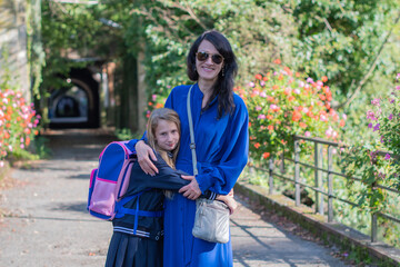 Fototapeta premium Young brunette in blue dress accompanies her little daughter to school, they walk together in the park, and the girl lovingly hugs her mother, back to school on a sunny day