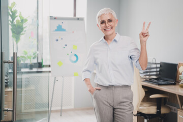 Photo of smiling positive mature lady accountant formalwear showing v-sign indoors workplace...