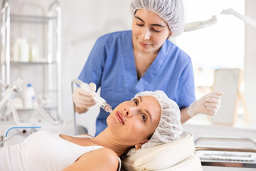Young female cosmetologist performs cleansing hardware facial procedure on young female patient