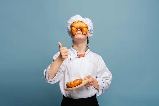 Teenager baker holding croissant on eyes showing thumbs up, fooling around