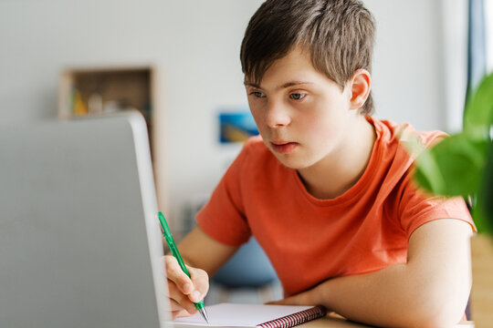 Focused teenager with down syndrome studying online, taking notes