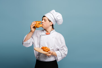 Baker smelling freshly baked pastry holding wooden tray standing isolated on blue background