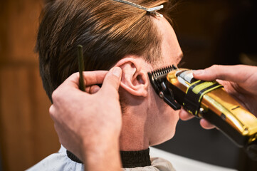 Close-up photo of professional barber shaving sideburns. Holding ear to make straight line. Competent hairdresser using electric shaver for work.