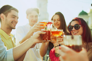 Smiling young adults, students enjoy sunny outdoor gathering, clinking glasses filled with colorful alcohol drinks. Concept of party, Friday mood, celebration, summer holidays, relax.