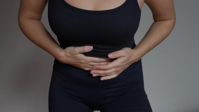 Cropped shot of a young woman holding her stomach with her hands on a beige background. Belly pain, abdominal distension, colic, digestive disorders, gastritis, stomach and duodenal ulcer