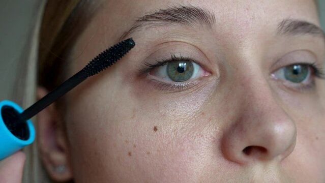Young caucasian blonde woman using a brush applying black mascara to the eyelashes on a gray background. The result of lengthening and increasing the volume of natural lashes