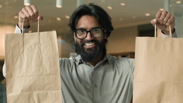 Happy joyful indian man with glasses indoors male buyer client guy customer shopper looking camera smiling showing paper bags satisfied purchases discount sale in store shows purchase goods packages