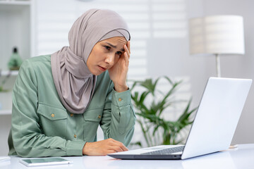 Businesswoman wearing hijab feeling stressed while working on laptop in modern office. Concept of work stress, fatigue, remote work challenges, and concentration.