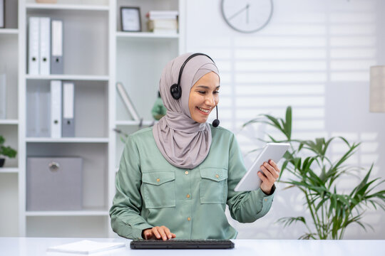 Smiling businesswoman in hijab using tablet for remote work in modern office. Woman wearing headset working remotely, engaging in video call or online meeting. Business concept