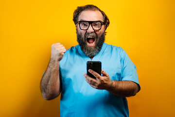 Euphoric doctor in blue coat on yellow background using smartphone. Concept of celebrating a victory by raising your fist.