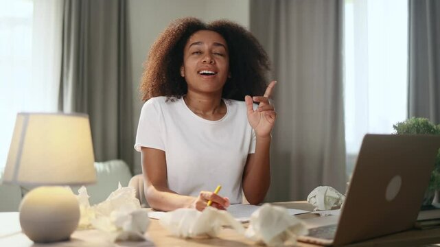 Portrait of young curly woman student or freelancer thinks questioningly and finding solution and writing on paper at home workplace Idea pointing forward index finger concept