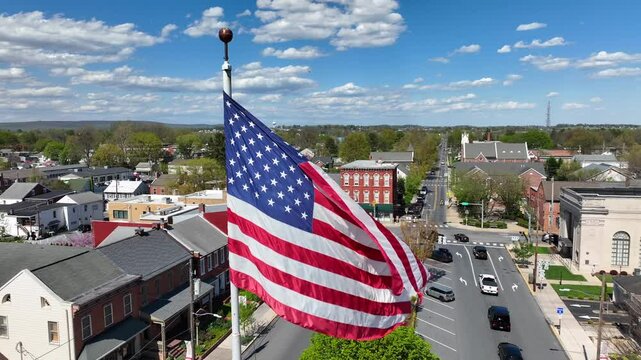 Patriotic American flag waves over small town in USA.  Main Street in Small Town America square at sunny day in spring. Aerial close up orbit shot.