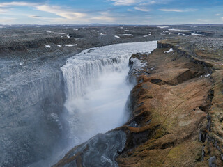 Stunning aerial view of a powerful waterfall in Iceland, surrounded by a barren rocky landscape with patches of green vegetation. Captures Iceland's dramatic beauty.